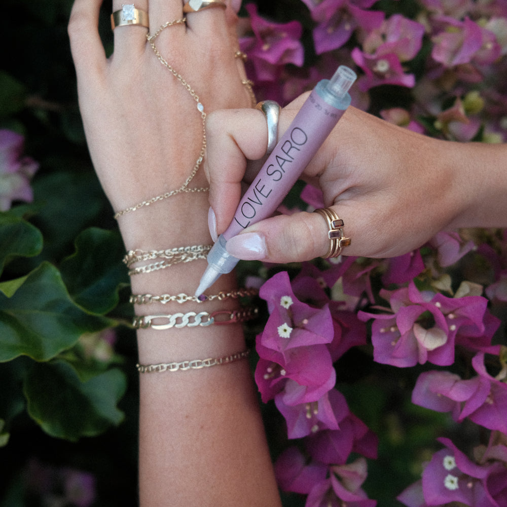 A wrist adorned with custom love saro permanent jewelry is using a cleaning pen to care for the 14K solid gold bracelets