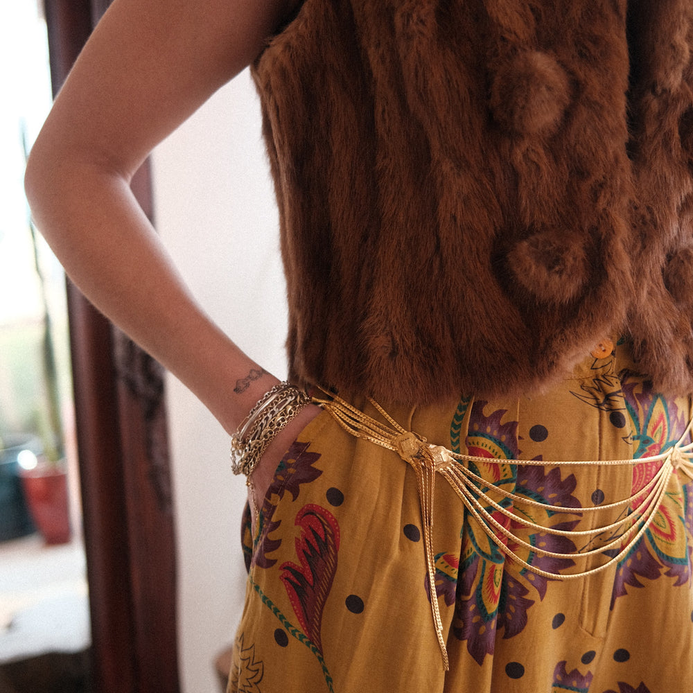 A woman in a yellow dress and fur vests models a vintage Christian Dior gold waist best chain belt wearing various gold custom bracelets
