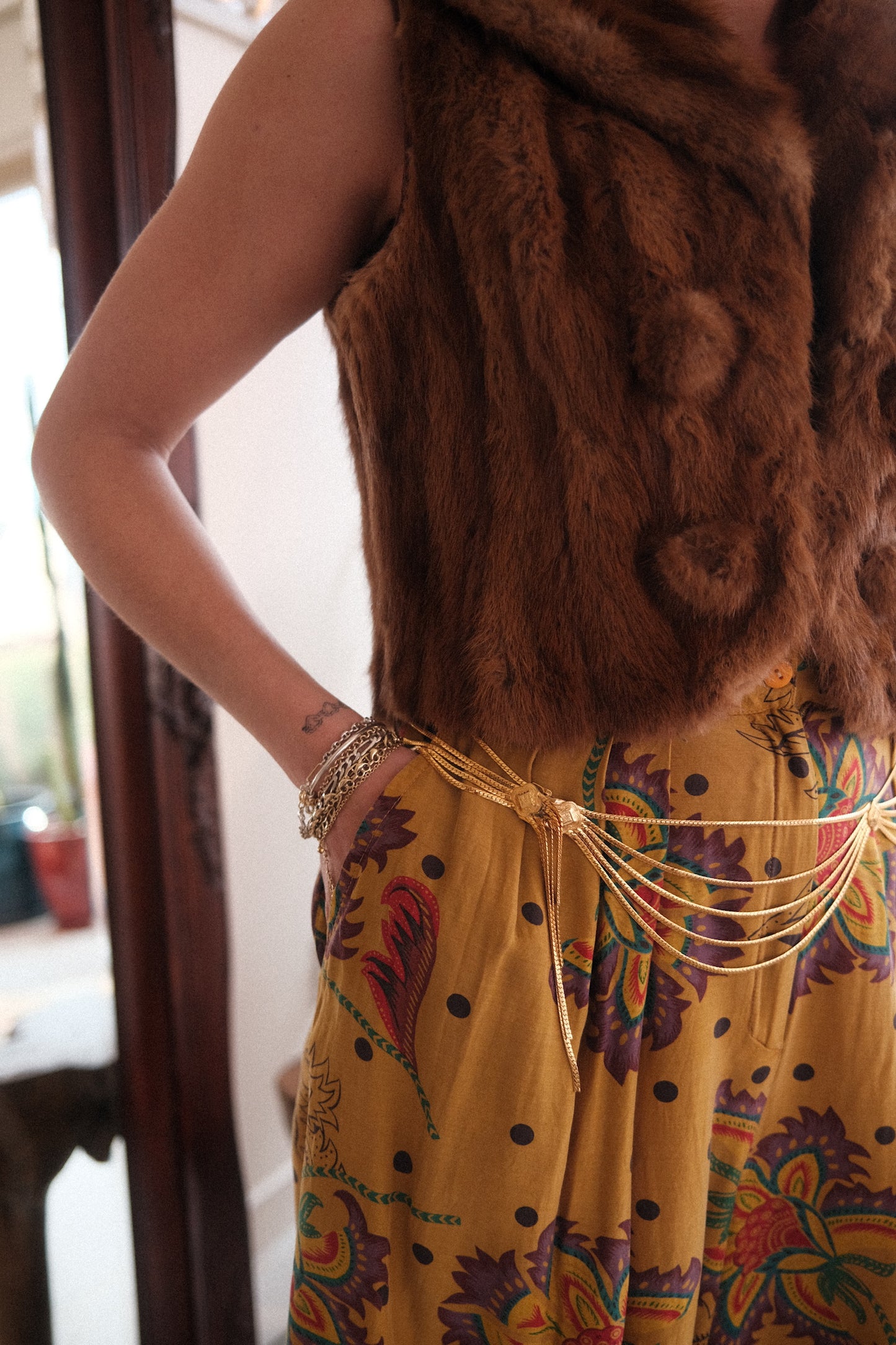 A woman in a yellow dress and fur vests models a vintage Christian Dior gold waist best chain belt wearing various gold custom bracelets